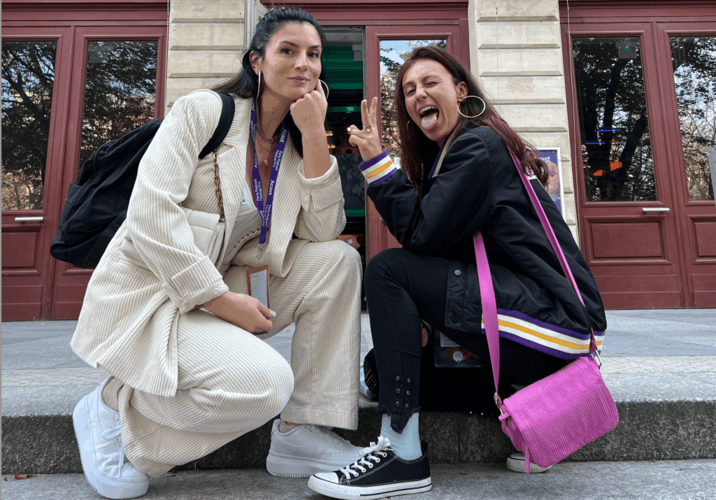 Chloé et Anouck posent devant un bâtiment, l'une en tailleur beige et l'autre avec un sac rose vif, souriantes.